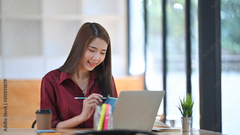 photo of young beautiful designer in red shirt taking notes in front of her laptop at the wooden desk over the modern office background. photo of young beautiful designer in red shirt taking notes in front of her laptop at the wooden desk over the modern office background.