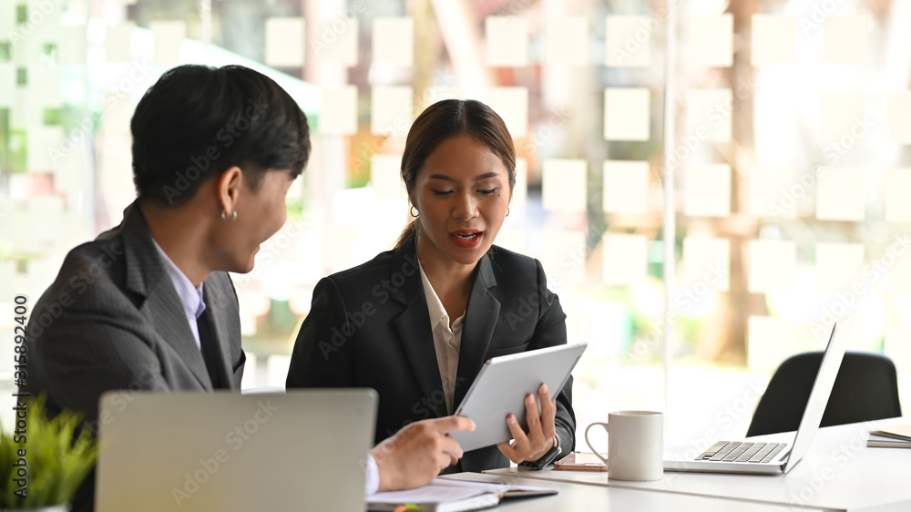 Photo of young confident business woman explaining a new project to her colleague next to her while sitting in the meeting room at the modern office. Photo of young confident business woman explaining a new project to her colleague next to her while sitting in the meeting room at the modern office.