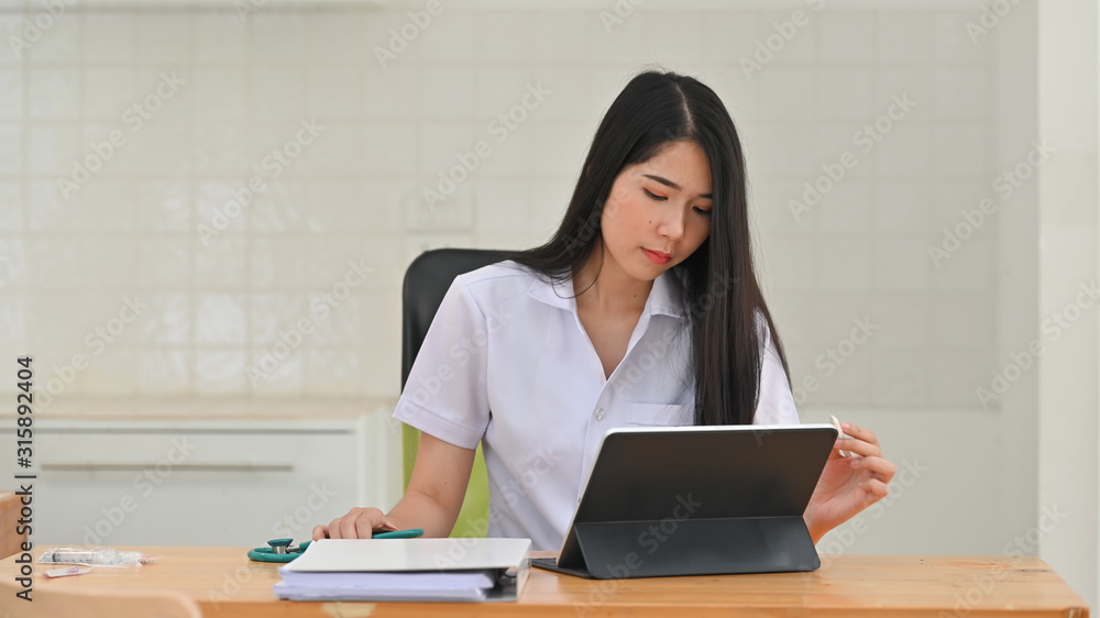 Photo of beautiful woman doctor in white uniform using tablet while sitting at work desk in hospital. Working young female medical staff concept. Photo of beautiful woman doctor in white uniform using tablet while sitting at work desk in hospital. Working young female medical staff concept.