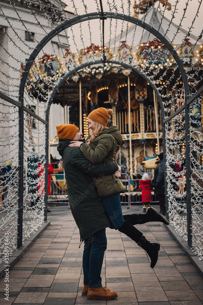 Beautiful cheerful couple in orange hats hugging and kissing on a blurry background with lights. He and she walking together on winter holidays. Valentines day concept.