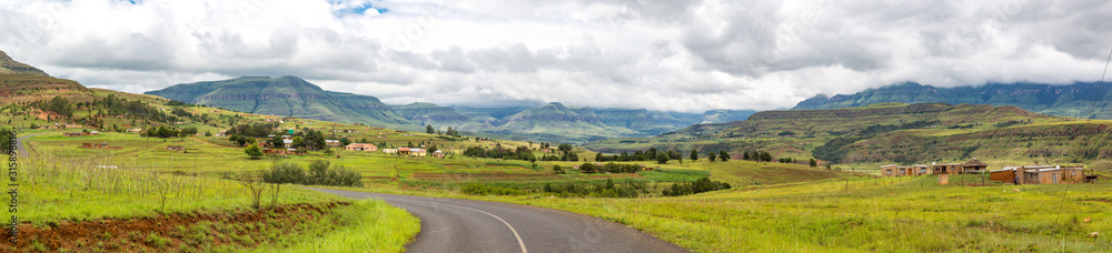 Panorama with a street leading to the mountains of Maloti Drakensberg, little village at the roadside, cloudy day, South Africa