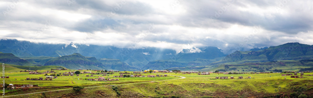 Panorama of a village in the mountains of Maloti Drakensberg Park with sunbeams breaking through the clouds and illuminating the green meadows, South Africa
