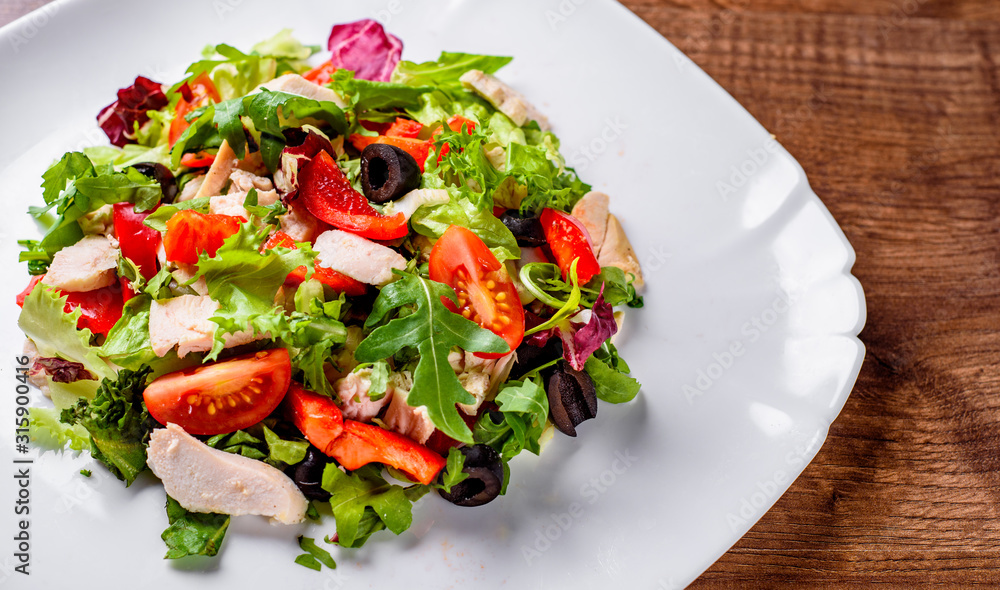 Fresh salad with chicken breast, arugula, black olives,red pepper, lettuce, fresh sald leaves and tomato on a white plate on wooden table background Fresh salad with chicken breast, arugula, black olives,red pepper, lettuce, fresh sald leaves and tomato on a white plate on wooden table background