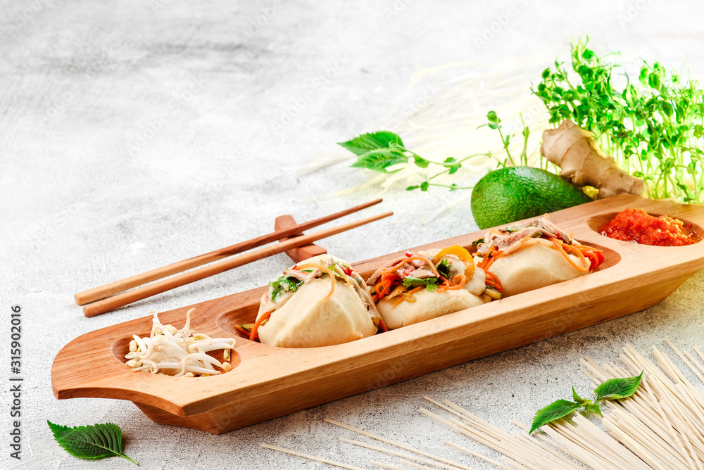 Bao - steamed pie with meat filling and fresh vegetables. Bao on beautiful wooden plate with chinese chopsticks, on light concrete background, decorated with fresh vegetables. Traditional chinese food