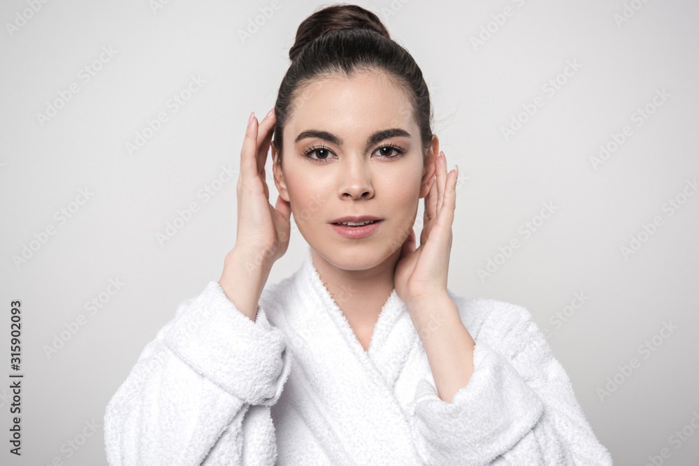 beautiful woman in bathrobe touching face while looking at camera isolated on grey beautiful woman in bathrobe touching face while looking at camera isolated on grey