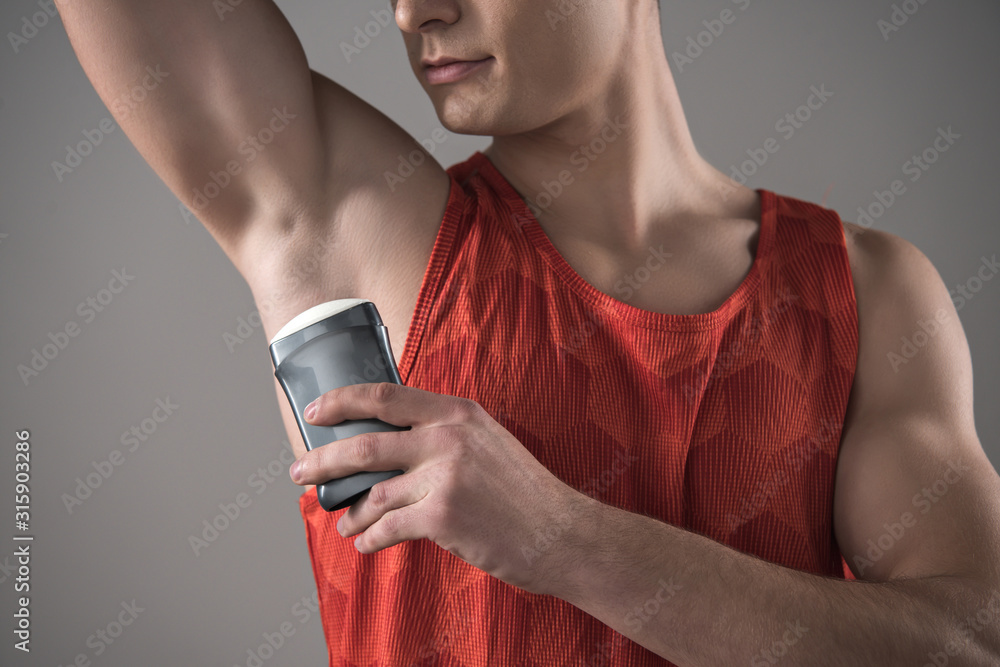 cropped view of young man in red sleeveless shirt applying deodorant on underarm isolated on grey cropped view of young man in red sleeveless shirt applying deodorant on underarm isolated on grey