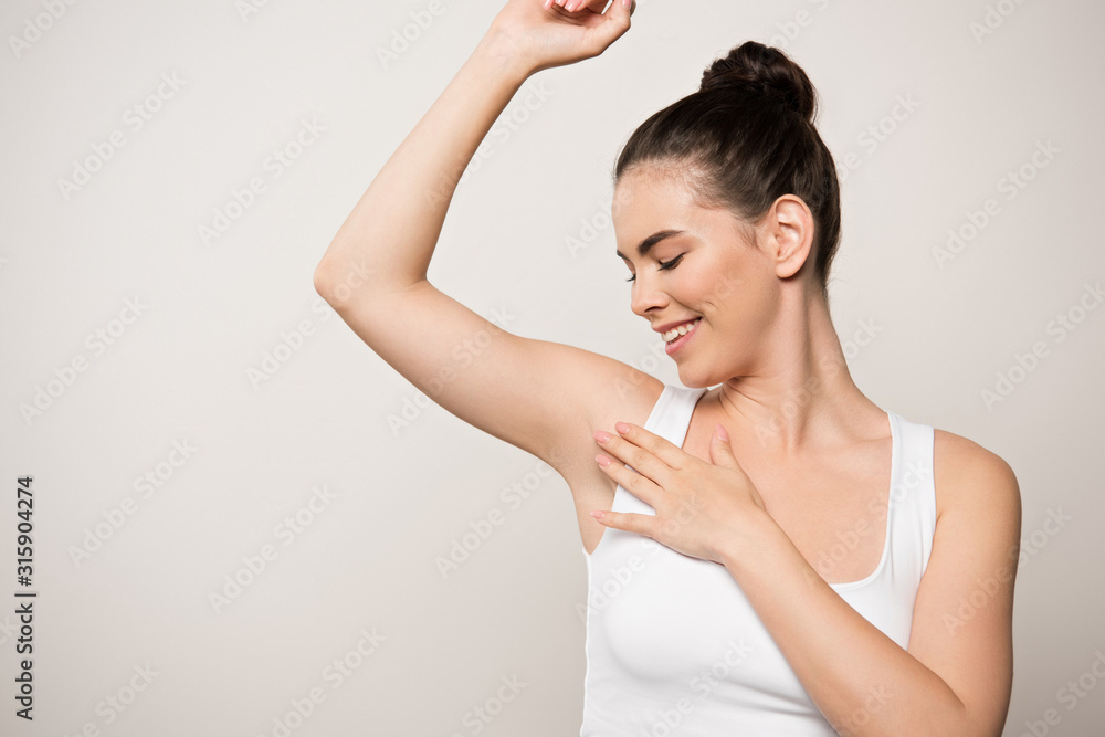 happy woman in white sleeveless shirt touching underarm isolated on grey happy woman in white sleeveless shirt touching underarm isolated on grey