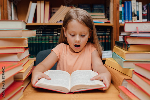 amazed little girl, looks in the book, is in the library, book background, indoor photo