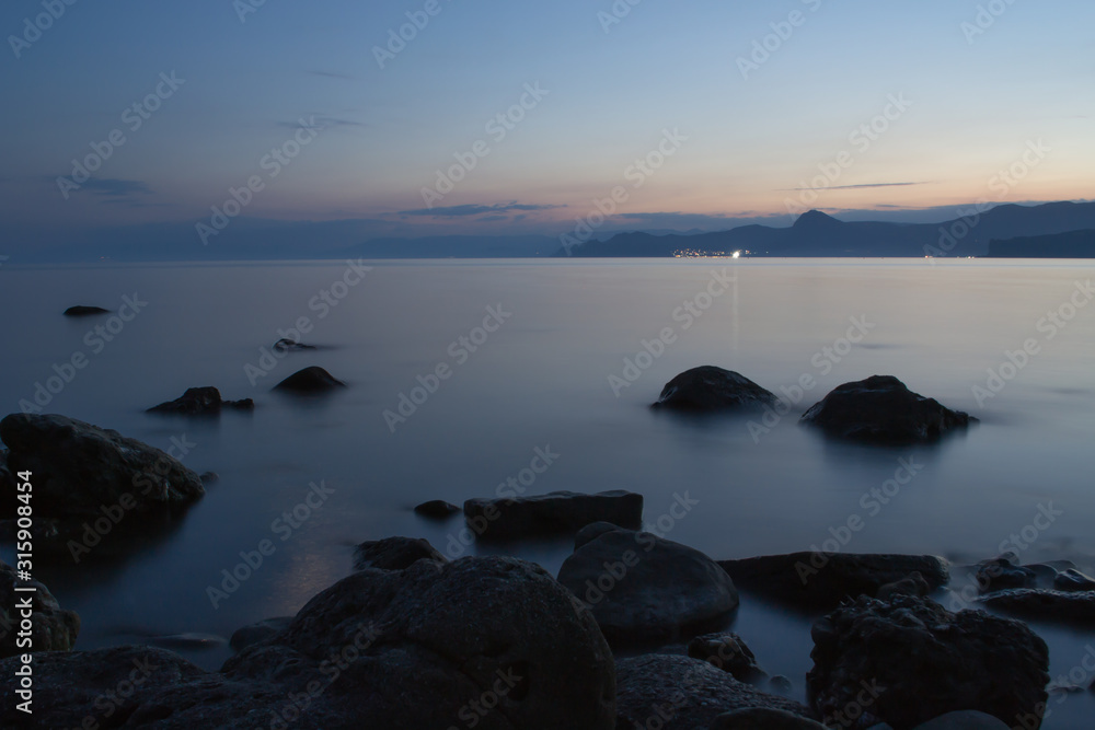 Fototapeta premium Sea at dusk. Stones on the shore and mountains in the distance. Sea and mountains after sunset. Photo in the dark. Fog on the water.