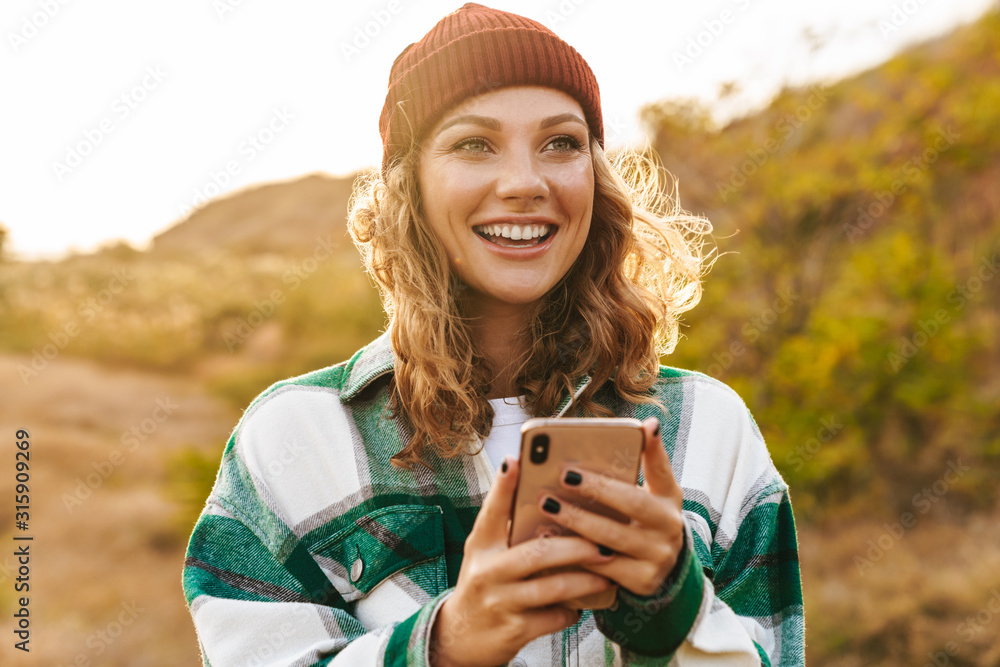 Image of joyful young woman holding cellphone while walking outdoors - obrazy, fototapety, plakaty 