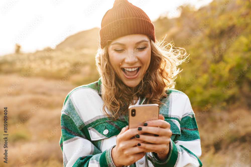 Image of joyful young woman holding cellphone while walking outdoors - obrazy, fototapety, plakaty 