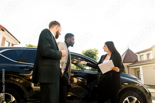 Young business people, two men, Caucasian and African, standing at the yard of car salon outdoors with young woman salesperson or manager buying or renting new black car crossover