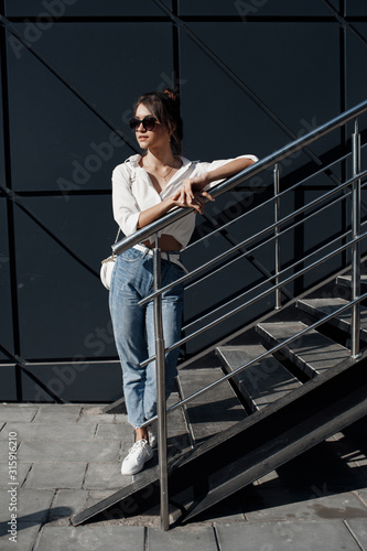A brunette woman with long hair in fashionable in blue jeans and a white shirt posing outdoors.Fashion photo concept. Woman near business center.Beautiful female business center worker near her office