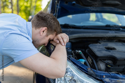 A sad man bowed his head over a broken car with an open hood on the side of the road. A man in despair next to his car.