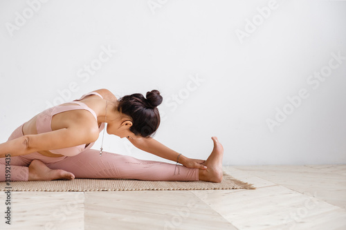 Close-up of a pacified young well-groomed brunette woman doing stretching while doing Pilates exercises sitting on a rug in a pink gymnastic suit on a white background. Place for advertising photo