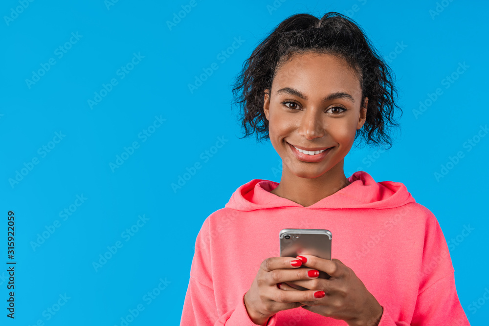 Smiling african young woman standing and using cell phone over blue background Smiling african young woman standing and using cell phone over blue background