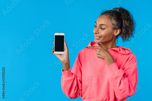Cheerful young afro woman holding mobile phone and pointing finger over blue background