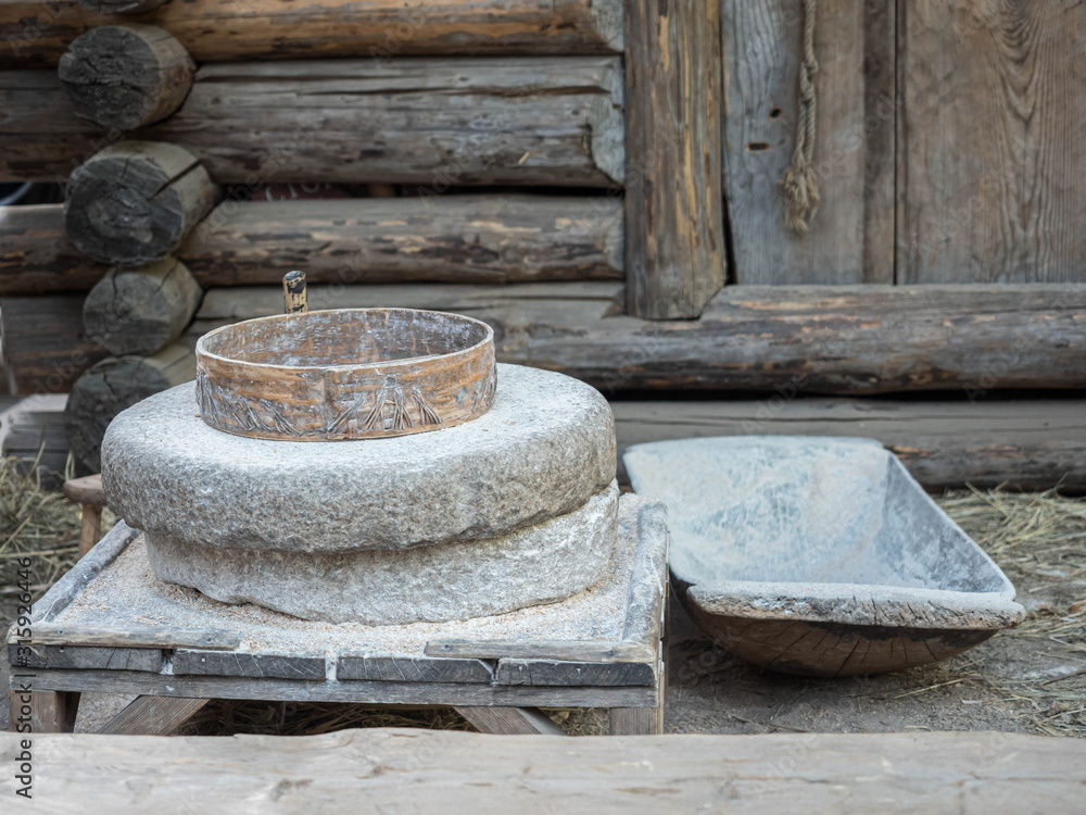Rotary discoid mill stone for hand-grinding a grain into flour. Medieval hand-driven millstone grinding wheat. The ancient Quern stone hand mill with grain near log house or russian izba