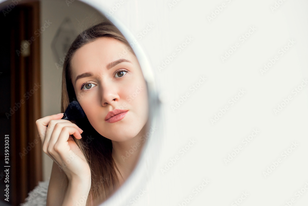 A young girl of Caucasian appearance looks in a round mirror and applies makeup, blush with a brush. A girl sits in a room and applies her daily face care and daily makeup. A young girl of Caucasian appearance looks in a round mirror and applies makeup, blush with a brush. A girl sits in a room and applies her daily face care and daily makeup.