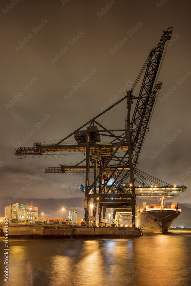 Night scene with illuminated container terminal, massive crane and moored vessel, Port of Antwerp, Belgium.