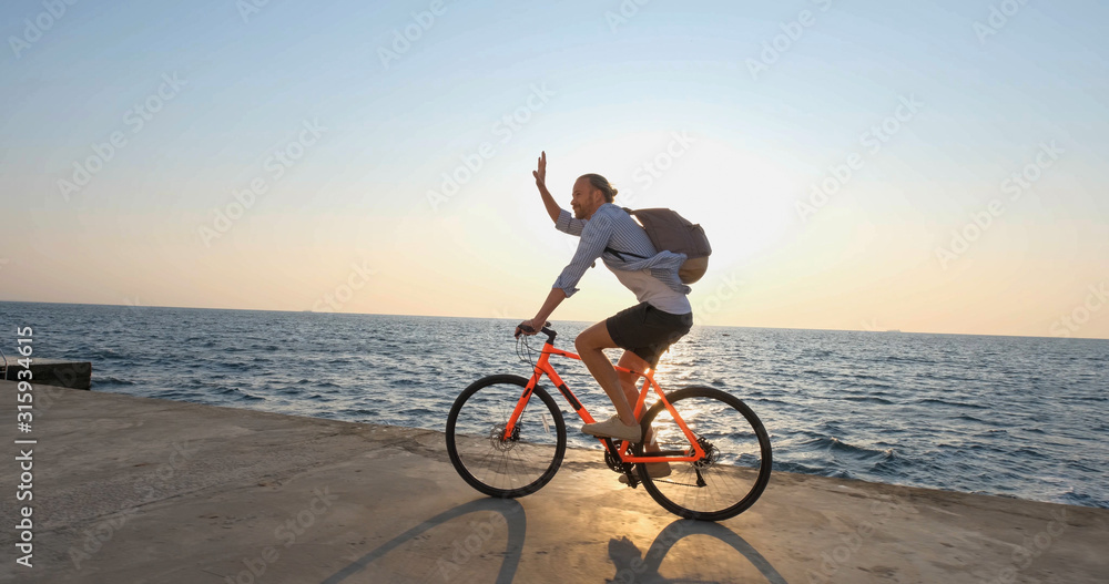 Fototapeta premium Young handsome male in casual wear ride on the colorful bicycle on the morning beach against beautiful sunset and the sea