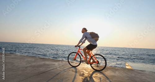 Young handsome male in casual wear ride on the colorful bicycle on the morning beach against beautiful sunset and the sea