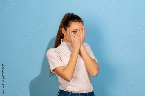 Covering face by hands. Caucasian teen girl's portrait on blue background. Beautiful model in casual wear. Concept of human emotions, facial expression, sales, ad. Copyspace. Looks scared, unhappy.