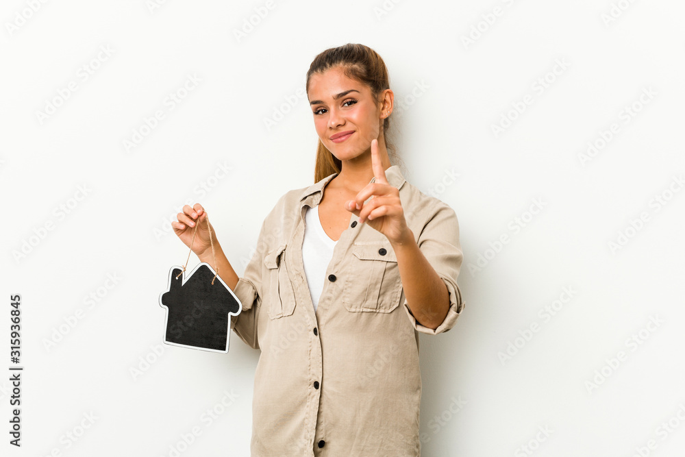 Young caucasian woman holding a house icon showing number one with finger. Young caucasian woman holding a house icon showing number one with finger.