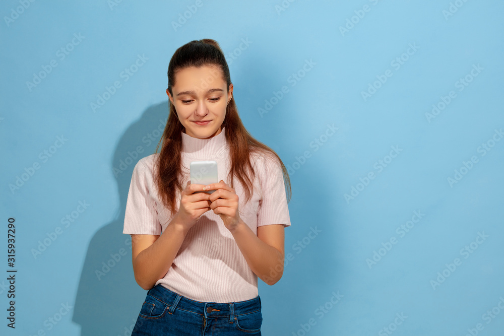 Smiling, using smartphone. Caucasian teen girl's portrait on blue background. Beautiful model in casual wear. Concept of human emotions, facial expression, sales, ad. Copyspace. Looks cute, happy. Smiling, using smartphone. Caucasian teen girl's portrait on blue background. Beautiful model in casual wear. Concept of human emotions, facial expression, sales, ad. Copyspace. Looks cute, happy.