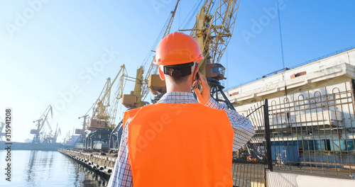 male worker of sea harbor in orange helmet and safety west, cranes and sea background  photo