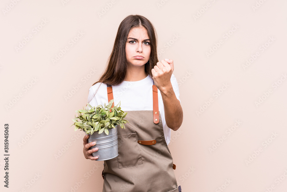 Young gardener caucasian woman holding a plant isolatedshowing fist to camera, aggressive facial expression. Young gardener caucasian woman holding a plant isolatedshowing fist to camera, aggressive facial expression.