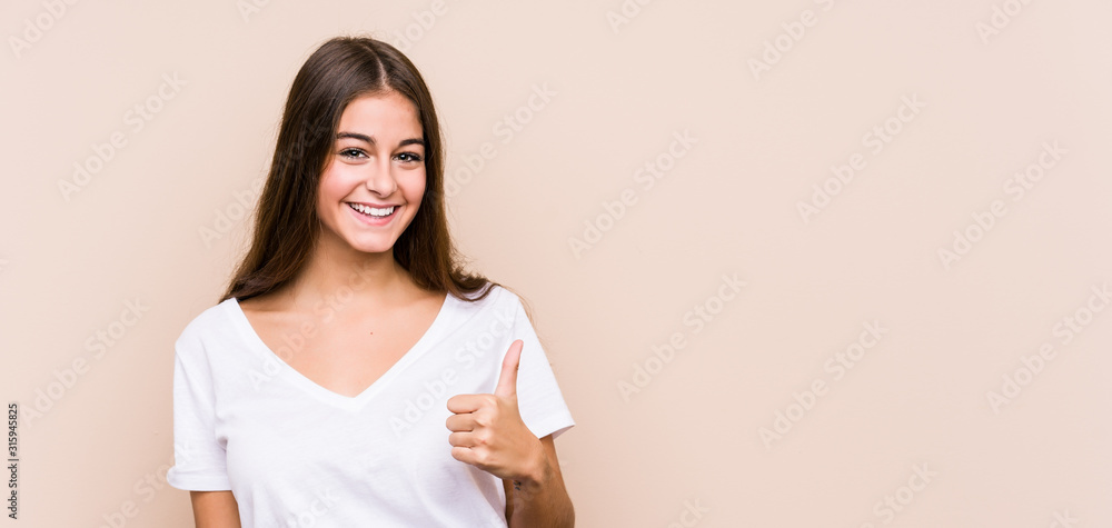 Plakat Młoda kaukaska kobieta pozuje na białym tle, uśmiechając się i podnosząc kciuk w górę