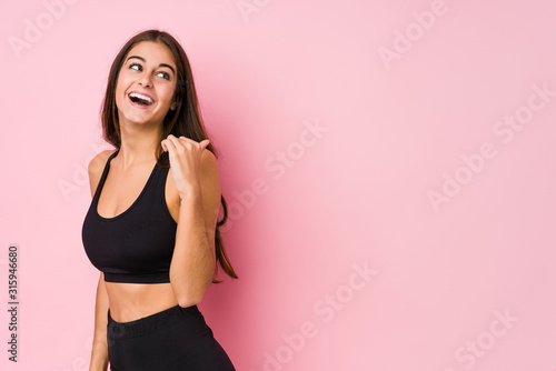 Young caucasian fitness woman doing sport isolated points with thumb finger away, laughing and carefree.