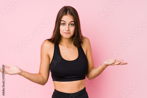Young caucasian fitness woman doing sport isolated doubting and shrugging shoulders in questioning gesture. © Asier