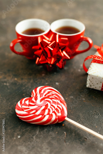 Two red cups of espresso or coffee, beautiful wrapped gift and decorative bow on a dark textured background. White-red lollipop in the form of a heart close up. Valentines day romantic date concept