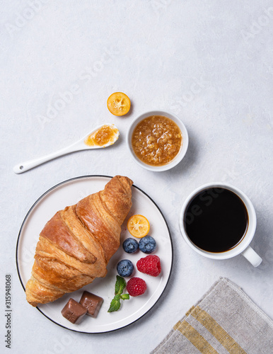 Fresh croissant on a white plate with kumquat jam, blueberries and raspberries with a Cup of vegan coffee on a gray background.