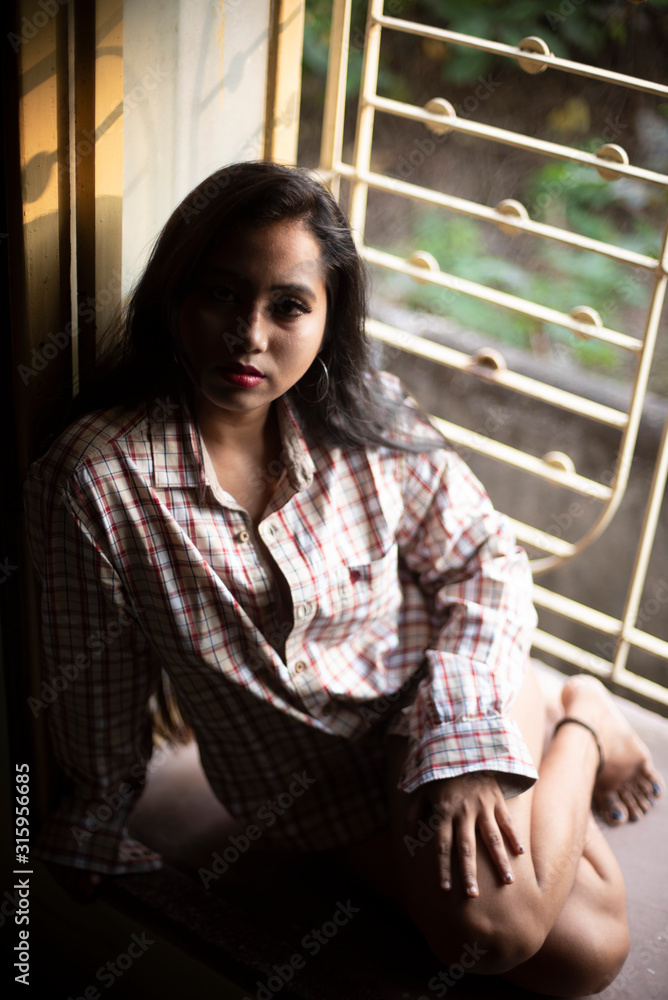 An young and attractive Indian  brunette woman in long western checkered shirt sitting on a window/balcony. Indian lifestyle.
