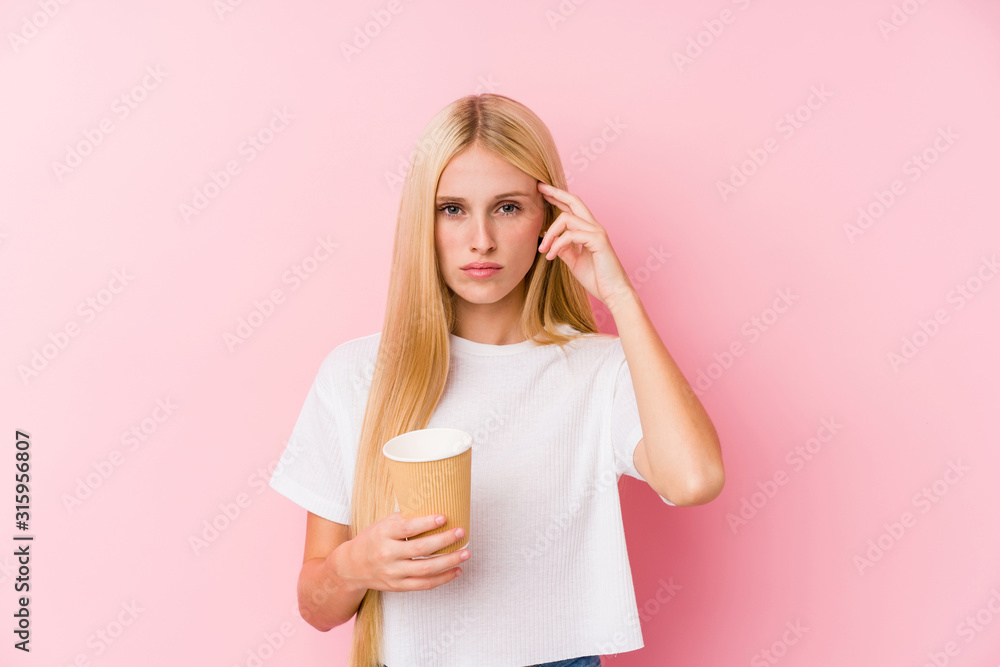 Young blonde girl holding a takeaway coffee pointing his temple with finger, thinking, focused on a task. Young blonde girl holding a takeaway coffee pointing his temple with finger, thinking, focused on a task.