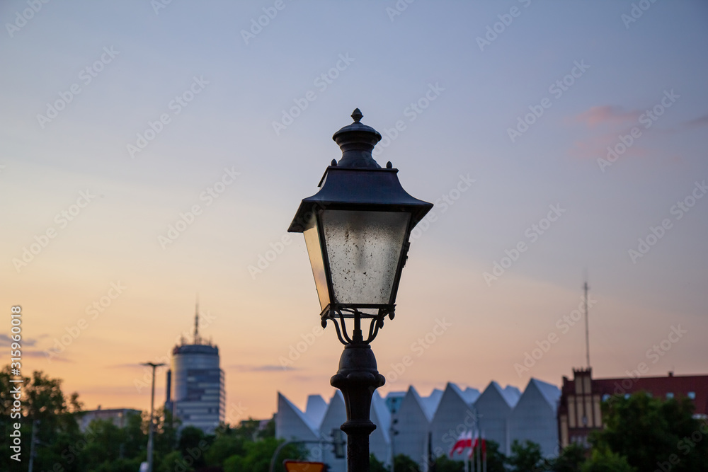 An old style lamp post in focus with the rest of town not in focus in the background as the sun sets over the city of Stettin, Poland