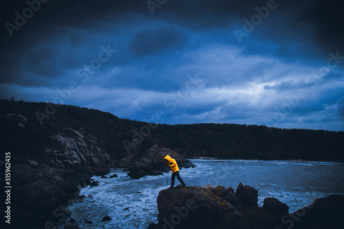 A young man adventurer with contrast yellow jacket looking down from a cliff by sea under gloomy sky with dramatic clouds