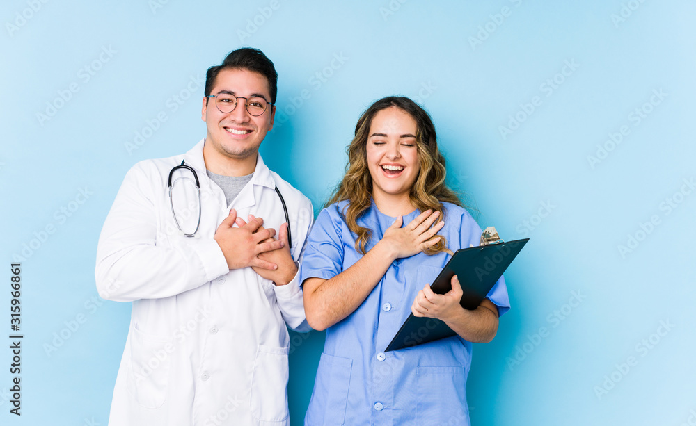 Young doctor couple posing in a blue background isolated laughing keeping hands on heart, concept of happiness. Young doctor couple posing in a blue background isolated laughing keeping hands on heart, concept of happiness.