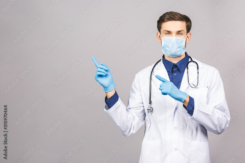 Potrait of handsome doctor wearing surgical protective mask and blue gloves isolated on grey background. Pointing finger.