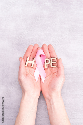 healthcare and medicine concept - female hands holding pink breast cancer awareness ribbon and letters of the word HOPE on gray background