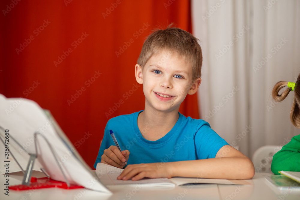smiling pretty fair-haired blue-eyed boy 9, a schoolboy, years old does homework, homework at the table, red background