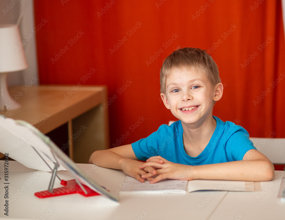 smiling pretty fair-haired blue-eyed boy 9, a schoolboy, years old does homework, homework at the table, red background smiling pretty fair-haired blue-eyed boy 9, a schoolboy, years old does homework, homework at the table, red background