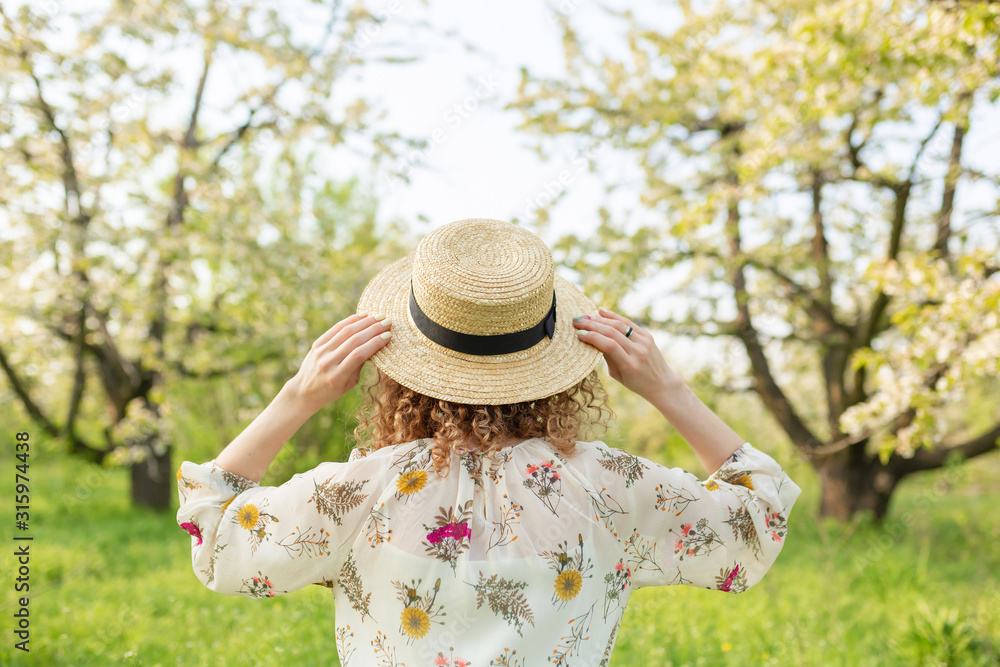 Rear view of a young woman with curly hair puts on a stylish wicker hat while walking in a green flowered garden. Spring mood