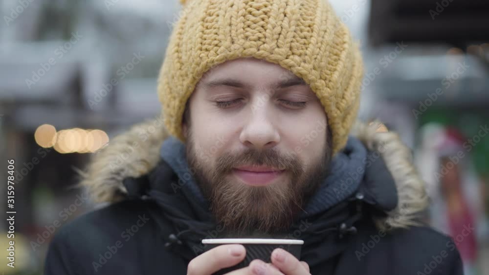 Close-up face of bearded Caucasian guy in yellow hat enjoying hot coffee outdoors. Young handsome man with brown eyes spending free time on winter fair. Lifestyle, happiness, leisure. Close-up face of bearded Caucasian guy in yellow hat enjoying hot coffee outdoors. Young handsome man with brown eyes spending free time on winter fair. Lifestyle, happiness, leisure.