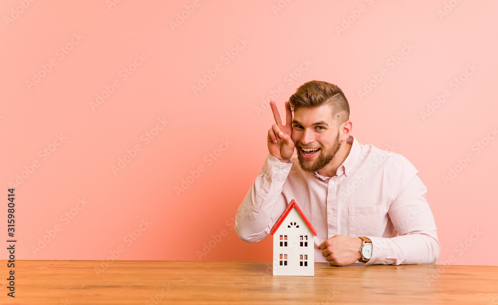 Young caucasian man sitting with a house icon showing victory sign and smiling broadly. Young caucasian man sitting with a house icon showing victory sign and smiling broadly.