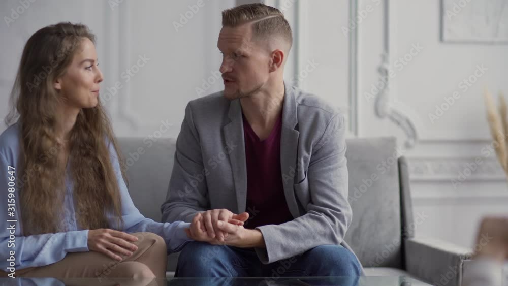 Tracking left of adult couple sitting on sofa holding hands and discussing family problems with professional therapist Tracking left of adult couple sitting on sofa holding hands and discussing family problems with professional therapist