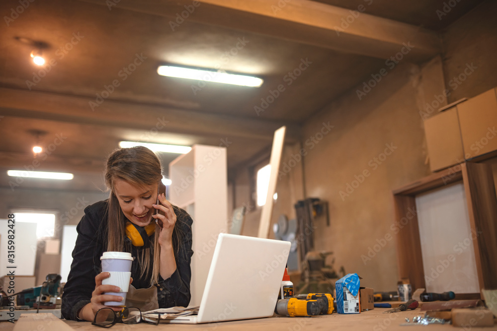 Young woman worker in the carpenter workroom. Woman making notes standing at table in modern workshop, copy space. Good looking young confident woman working as carpenter in her own woodshop Young woman worker in the carpenter workroom. Woman making notes standing at table in modern workshop, copy space. Good looking young confident woman working as carpenter in her own woodshop